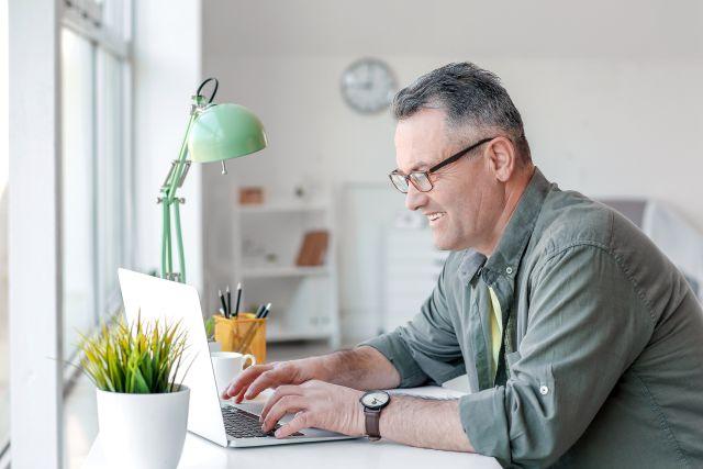 Man sitting at a desk by a window, smiling as he types on a laptop in a bright home workspace.