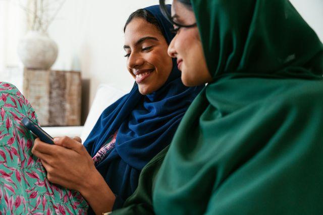 Two women wearing headscarves sitting together indoors, smiling as they look at a smartphone screen.