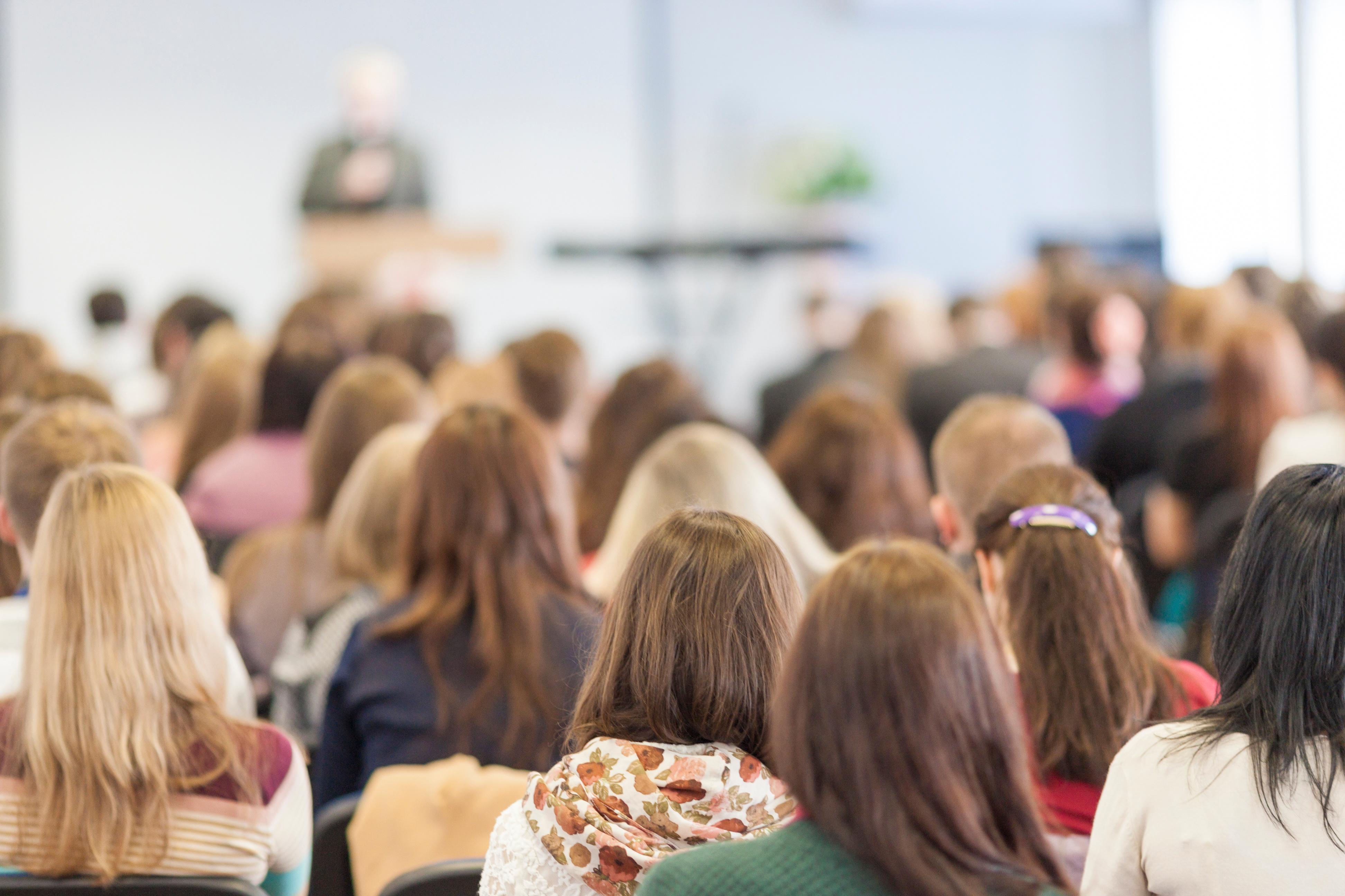 Audience seated indoors facing a speaker at the front of a room, with rows of people listening to a talk or presentation.