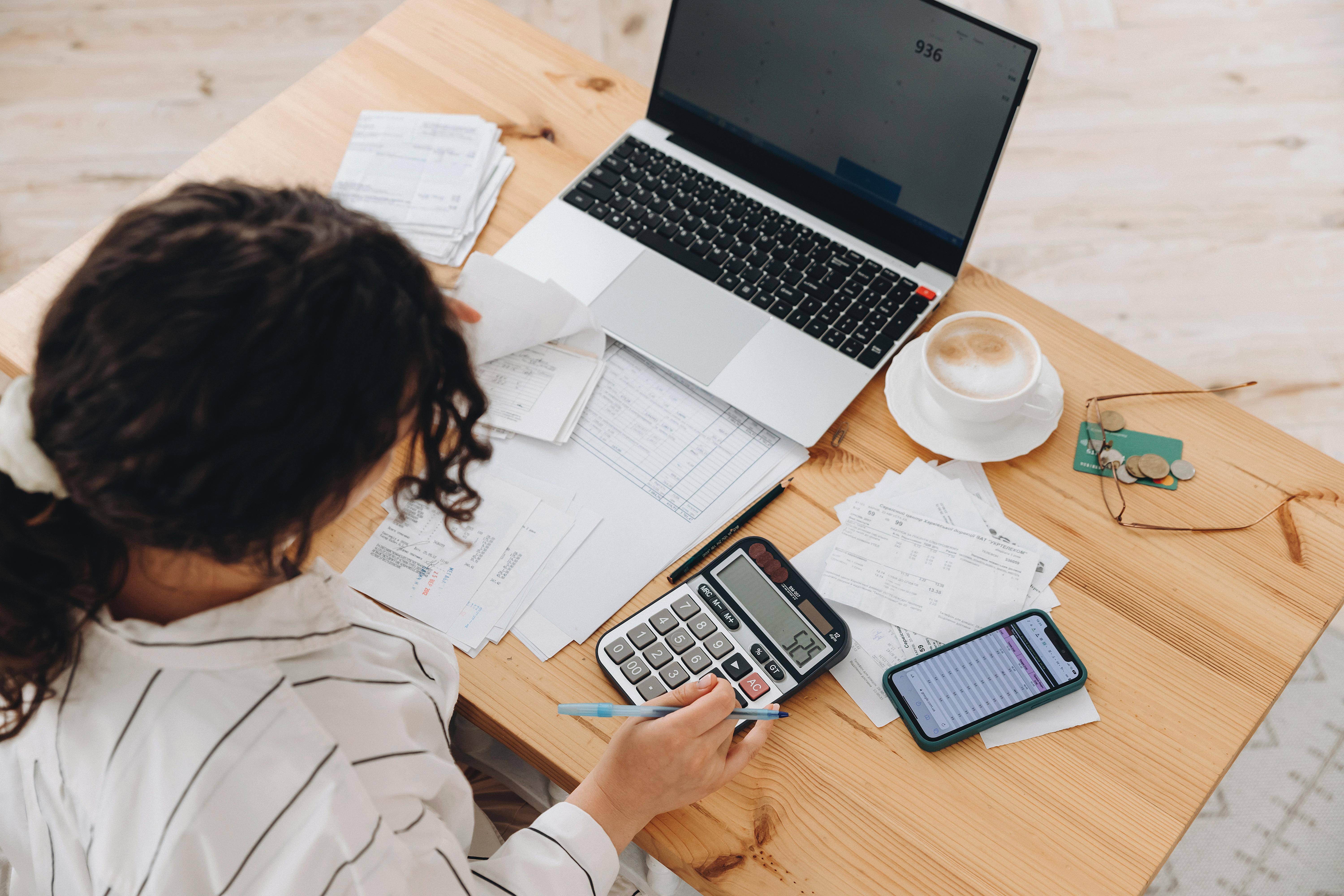 An overhead view of a person sitting at a wooden table, working on paperwork and calculations. A laptop is open in front of them, surrounded by scattered bills, receipts, and documents. The person holds a pen near a large calculator, with a smartphone displaying a spreadsheet beside it. A cup of coffee, a pair of glasses, and small coins rest on the table, suggesting budgeting or financial planning.