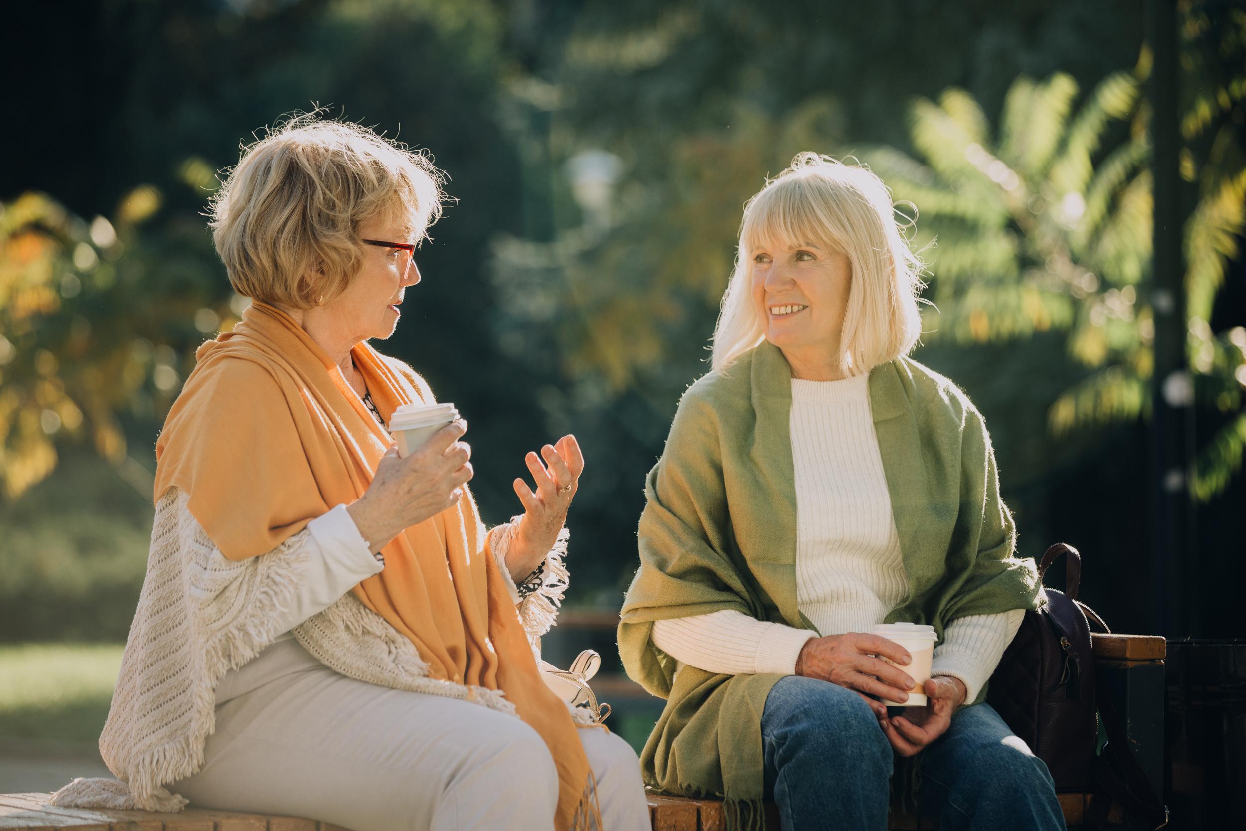 Two women sitting outdoors on a bench, holding takeaway cups and talking together in a green park setting.