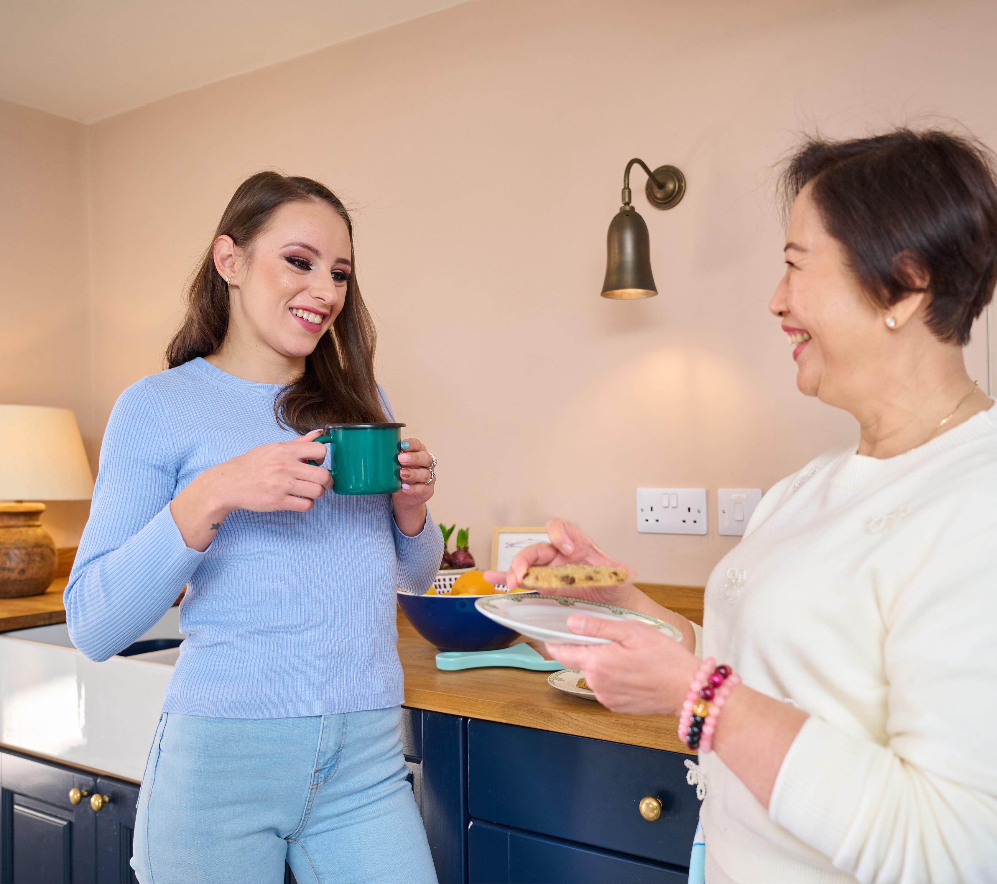Two women standing in a kitchen, smiling and talking together, one holding a mug and the other offering a plate of food.