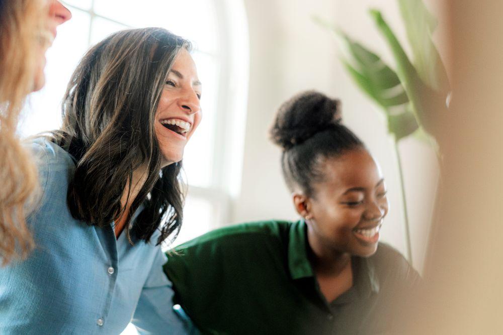 Two women indoors leaning together and laughing, sharing a light moment in a bright, welcoming space.