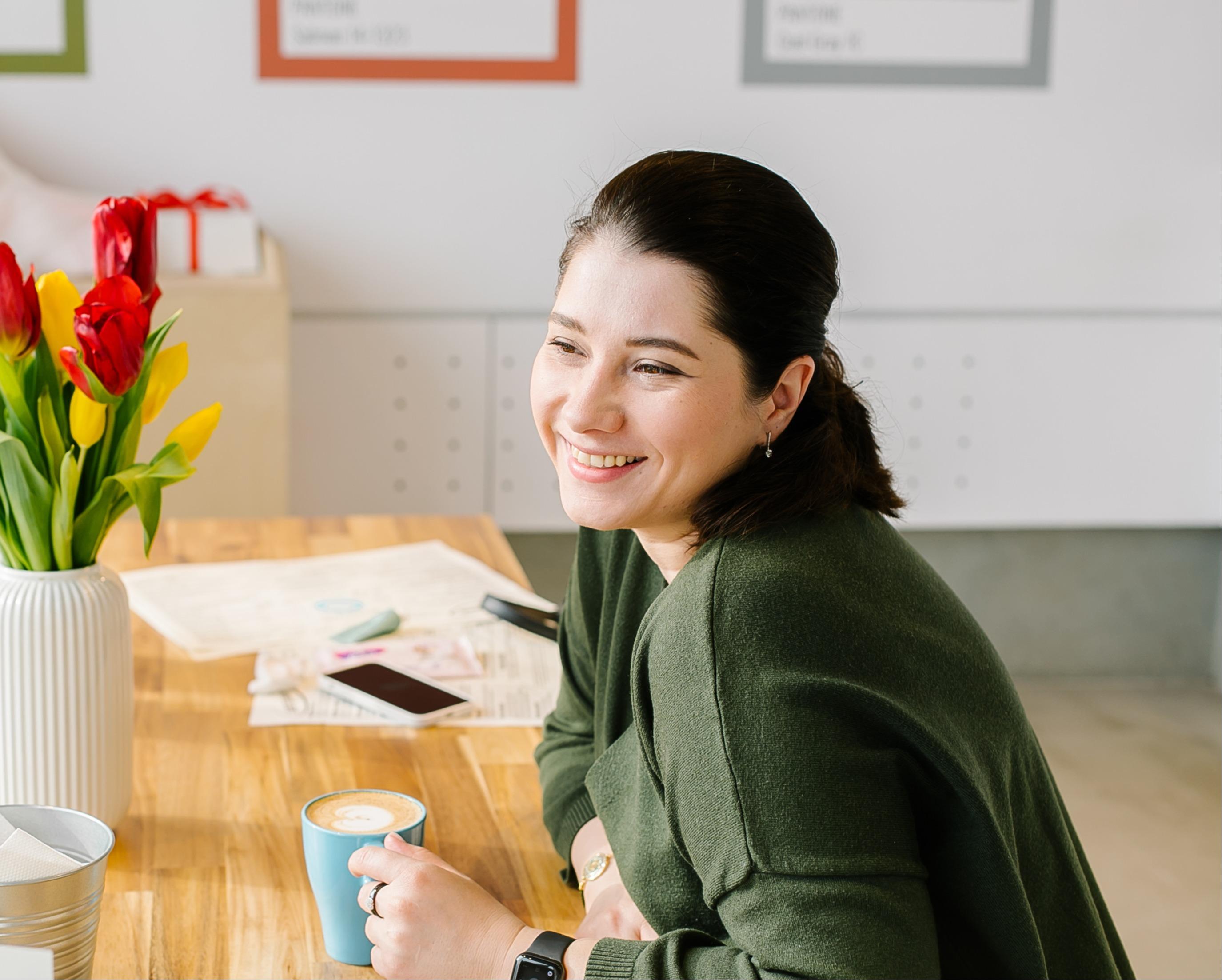A woman with dark hair pulled back smiles warmly while sitting at a wooden table in a bright indoor space. She wears a green sweater and holds a blue cup of coffee. On the table beside her are scattered papers, a smartphone, and a vase of red and yellow tulips. The background is softly out of focus, giving the scene a relaxed, friendly atmosphere.
